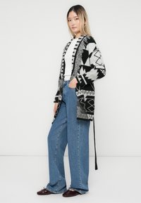Kvinne iført en svart og hvit geometrisk mønstret cardigan, hvit skjorte, vide blå jeans og mørkerøde flate sko, stående mot en hvit vegg.