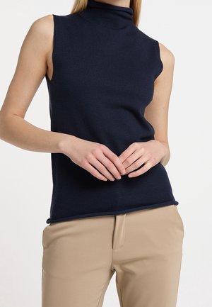 Mouwloze marineblauwe coltrui gedragen met beige broek, handen van het model rustig samengevouwen ter hoogte van de taille tegen een witte achtergrond.