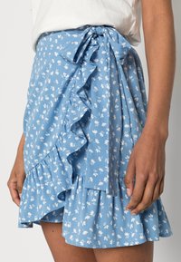 Jupe portefeuille bleue à motif floral blanc, ourlet à volants et nœud à la taille, confectionnée dans un tissu léger, présentant une texture douce.