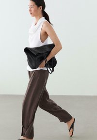 Frau läuft drinnen, trägt ein weißes ärmelloses Top, lockere braune Hose, schwarze Flip-Flops, trägt eine große schwarze geflochtene Tasche unter dem Arm.