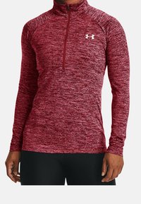 Uomo che indossa una maglietta a maniche lunghe color bordeaux melangiato con zip trimestrale aderente e logo bianco di Under Armour sul petto, abbinata a pantaloni neri.