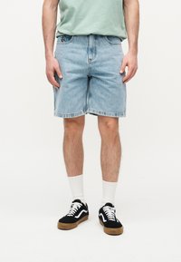 Άνδρας που φοράει κοντό ανοιχτό μπλε τζιν, μαύρα και λευκά αθλητικά παπούτσια, λευκές κάλτσες και ανοιχτό πράσινο πουκάμισο, στέκεται σε λευκό φόντο.