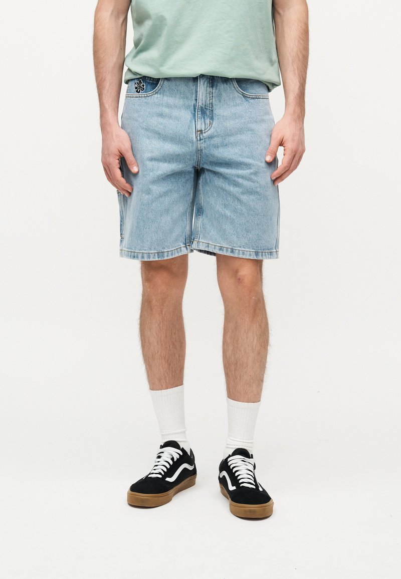 Άνδρας που φοράει κοντό ανοιχτό μπλε τζιν, μαύρα και λευκά αθλητικά παπούτσια, λευκές κάλτσες και ανοιχτό πράσινο πουκάμισο, στέκεται σε λευκό φόντο.