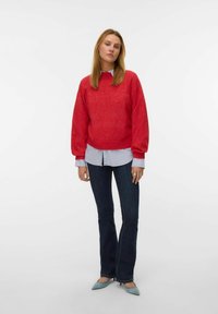 Suéter rojo de punto con mangas largas, superpuesto a una camisa de cuello azul claro. Combinado con jeans de campana oscuros y zapatos azulados de punta.