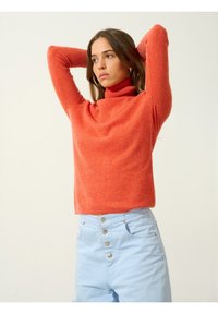 Roter Rollkragenpullover aus weichem, strukturiertem Stoff, kombiniert mit hellblauen, hochgeschnittenen Hosen mit silbernen Knopfakzenten.