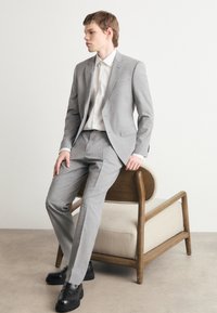 Completo grigio chiaro con un motivo testurizzato, vestibilità su misura, camicia bianca e scarpe eleganti nere, seduto su una sedia di legno con un cuscino di tessuto.