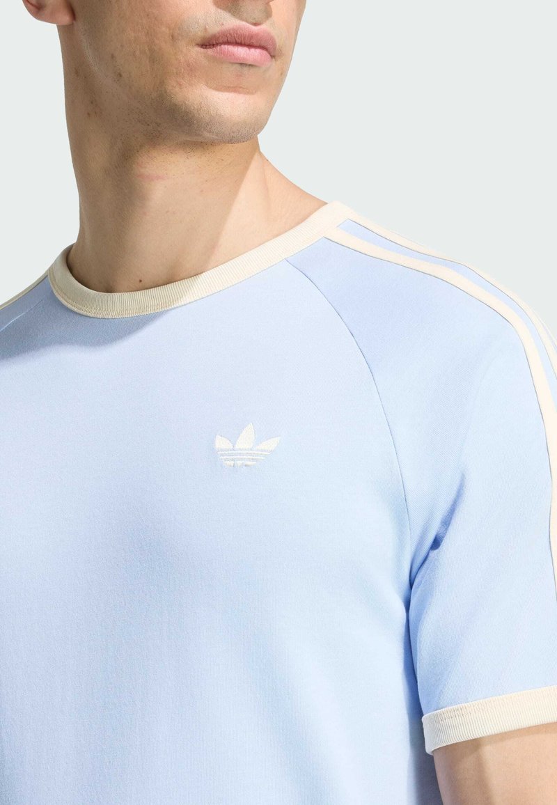 Uomo con maglietta Adidas azzurra con rifiniture beige e logo sul petto, che guarda di lato su uno sfondo uniforme.