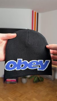 Zwarte gebreide muts met gedurfde, blauwe en witte letters "obey". Zacht van textuur en volgt de vorm van het hoofd, zonder zichtbare hardware of accenten.
