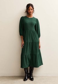 Grünes, gestuftes Kleid aus strukturiertem Stoff mit Puffärmeln und rundem Ausschnitt, kombiniert mit kniehohen schwarzen Stiefeln.