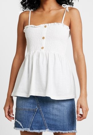 Mujer vestida con una blusa sin mangas de tirantes blancos con botones y una falda mini de mezclilla azul deshilachada, de pie con los brazos relajados a los lados.