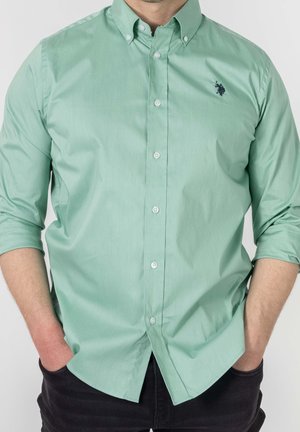 Homme portant une chemise boutonnée vert clair aux manches retroussées et un pantalon noir, les mains dans les poches, faisant face à l'avant.