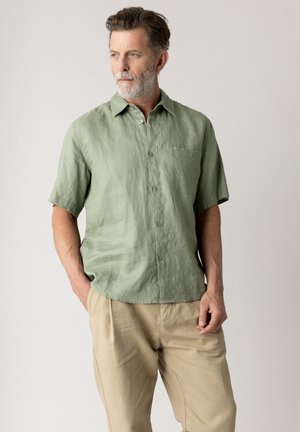 Mann mit grauem Bart, der ein hellgrünes Kurzarmhemd und beige Hose trägt, steht mit den Händen in den Taschen und schaut nach links.