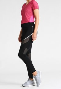 Červené sportovní tričko s růžovým bočním akcentem, v kombinaci s černými legínami s síťovinovými vsadkami a reflexním pruhem. Šedé tenisky dotvářejí celkový vzhled.