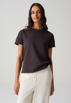 Schwarzes Baumwoll-T-Shirt mit aufgerollten Ärmeln, rundem Halsausschnitt und dezentem Logo auf der Brust, kombiniert mit hellbeigen, locker sitzenden Hosen.