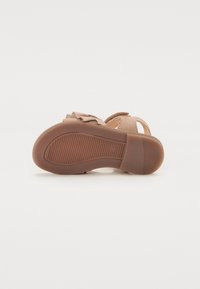 Ljusbrun sandal med platt sula, texturerad gummibotten och dekorativ rosett. Öppen bak och på sidorna för bättre andning.