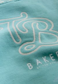 Jasnoniebieska tkanina z białym haftowanym wzorem i napisem "BAKED" w płynnej czcionce oraz teksturowanej powierzchni.