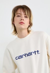 Cremefarvet sweatshirt med et marineblåt broderet "carhartt" logo på tværs af brystet. Har en rund hals og ribbede manchetter.