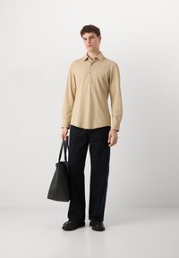 Camisa beige de manga larga y botones con tejido texturizado, combinada con pantalones azul oscuro. El modelo sostiene una bolsa tote negra.