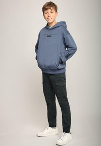 Felpa blu con grafica frontale, tasca e polsini a coste, abbinata a pantaloni cargo scuri e sneaker bianche. Texture complessivamente liscia.