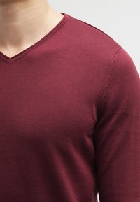 Burgundy V-kaula neule, jossa on teksturoitu neulonta, nähden tiukan muotoilun ja huomaamaton ommel yksityiskohtana olkatikkauksessa.