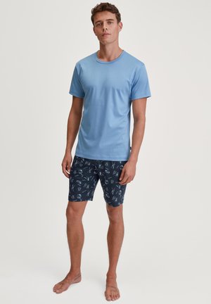 Jonge man die op blote voeten staat, gekleed in een lichtblauw T-shirt en marineblauwe korte broek met een wit en rood patroon, tegen een witte achtergrond.