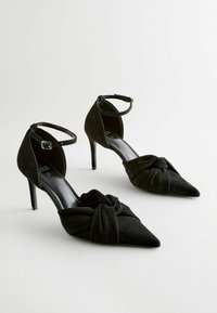 Musta mokkanahkainen korkokengät, joissa on terävät varpaat, solmittu yksityiskohta edessä ja nilkkahihnat. Tyylikäs muotoilu ja stilettokorko.