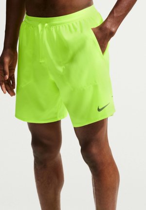 Homme portant un short de sport jaune néon vif avec un logo Nike noir, une main dans la poche, sur un fond uni clair.