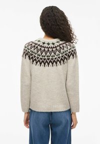 Osoba z długimi,kręconymi włosami, ubrana w beżowy sweter robiony na drutach z czarnym i zielonym wzorem oraz niebieskie dżinsy, widziana od tyłu.