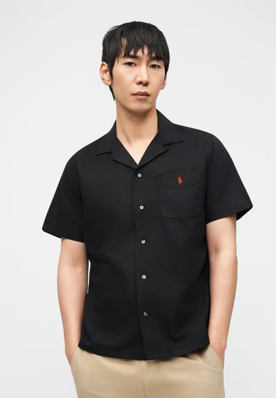 Άνδρας που φοράει μαύρο κοντομάνικο πουκάμισο με τσέπη στο στήθος και μπεζ παντελόνι, στέκεται με τα χέρια στις τσέπες σε λευκό φόντο.