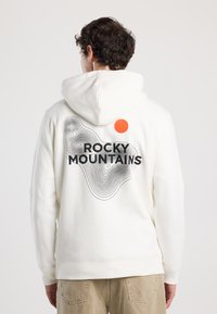 Felpa bianca con una grande grafica "ROCKY MOUNTAINS" in nero, caratterizzata da un motivo a linee ondulate e un accento circolare arancione nel colletto.
