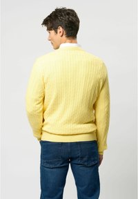 Gult kabelstickat tröja med ribbad nederkant, bärs över en vit skjorta med krage, parat med blå jeans, sedd bakifrån.