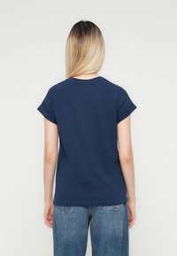 Frau mit langen, glatten, blonden Haaren, die ein kurzärmliges, dunkelblaues Hemd und Bluejeans trägt, steht vor einem einfarbigen hellen Hintergrund.
