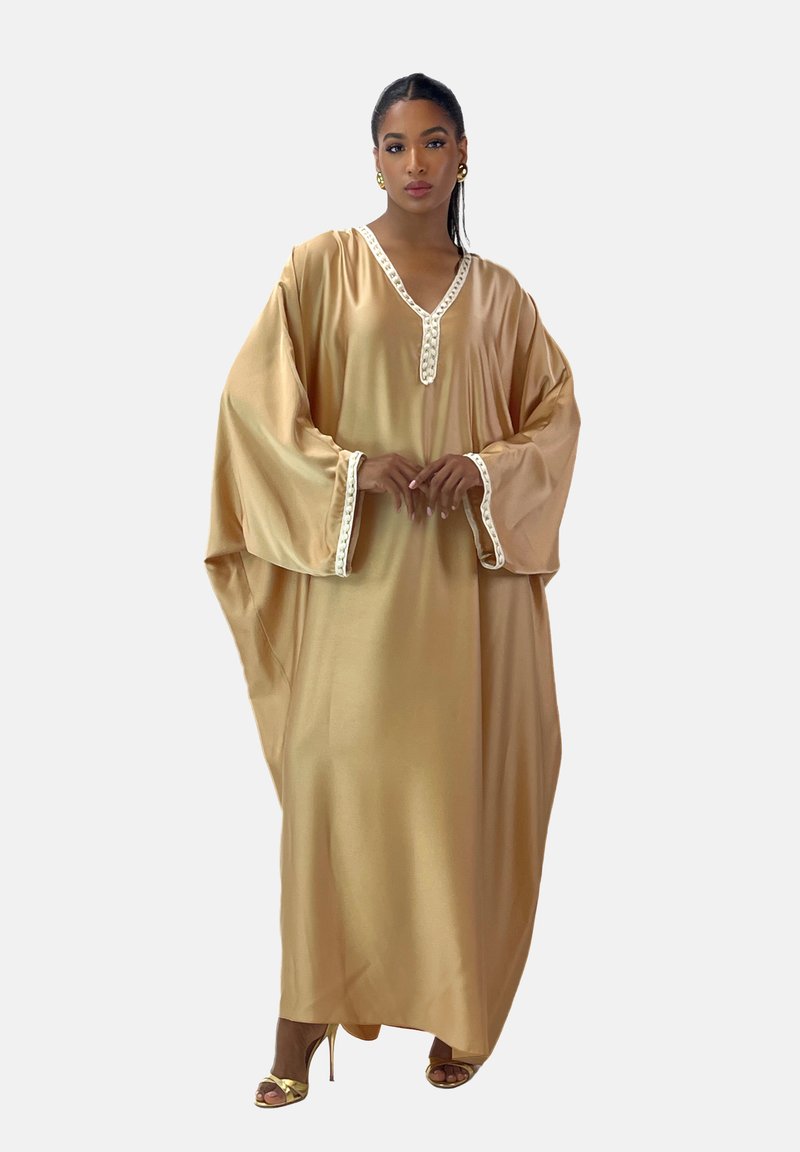 Satin kaftan i guld med V-hals og hvid kant, der har brede ærmer og et flydende design. Modellen bærer guld hæle med stropper.