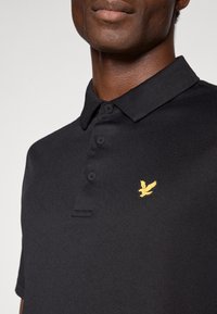 Melnā polo krekla, kas aprīkots ar trīs pogu aizdari, izšūtu dzeltenu ērgļa logo kreisajā krūtī un teksturētu audumu.