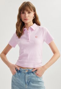 Nainen, jolla on pitkät hiukset, wearing vaaleanpunainen lyhythihainen polo-paita, jossa on pieni punainen sydän rinnassa, ja vaaleansiniset farkut, kädet taskuissa.