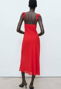 Robe midi rouge en tissu fluide avec des détails en volants sur les épaules et des bretelles réglables. Présente une texture légère et lisse.