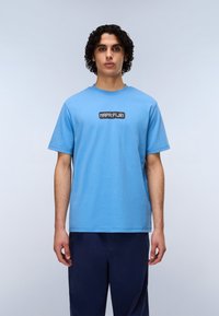 Ανοιχτό μπλε βαμβακερό μπλουζάκι με μαύρο ορθογώνιο λογότυπο και λευκό κείμενο μπροστά. Κοντά μανίκια και στρογγυλή λαιμόκοψη.