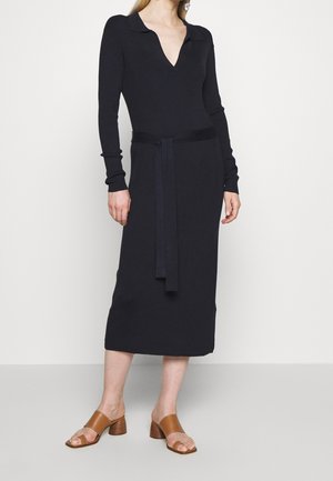 Femme portant une robe midi noire à manches longues avec col et ceinture, associée à des sandales à talons blocs ouvertes beige, debout devant un fond uni.