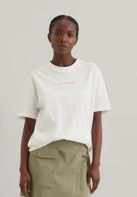 Hvid bomulds T-shirt med korte ærmer og rund hals, med orange tekst "Marcopolo Denim" på brystet. Stylet med en olivengrøn nederdel.