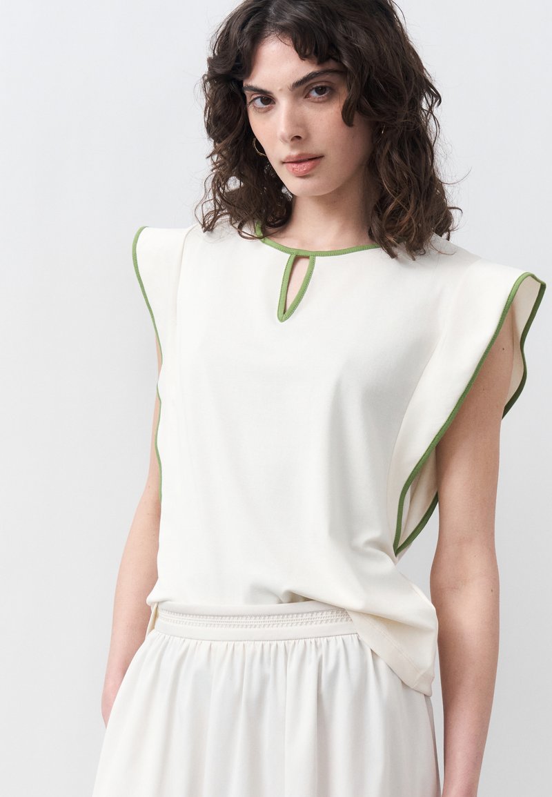 Γυναίκα με σκούρα σγουρά μαλλιά που φοράει λευκό αμάνικο τοπ με πράσινη λεπτομέρεια και άνοιγμα στο λαιμό, συνδυασμένο με λευκό φουστάνι με πτυχώσεις.