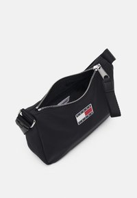 Borsa a tracolla nera in tessuto con zip argentata, tracolla regolabile e una patch con il logo con colori rosso, bianco e blu su uno sfondo rettangolare.