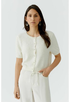 Frau mit langem dunklem Haar, die eine weiße, kurzärmelige, strukturierte Strickjacke mit Knöpfen und weiße Hosen mit Kordelzug trägt, steht mit einer Hand in der Tasche.