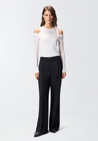 Witte lange mouwen top met uitsnijdingen bij de schouders, gecombineerd met hooggerende zwarte wijde broek en zwarte puntige schoenen.