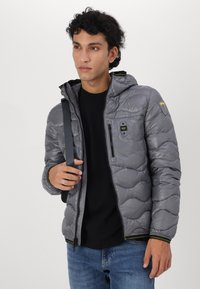 Graue Pufferjacke mit Kapuze, ausgestattet mit einer mit Reißverschluss versehenen Brusttasche, elastischen Bündchen und grünen Akzenten. Getragen über einem schwarzen Shirt mit Jeans.