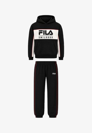 To unge voksne iført matchende sorte FILA hættetrøjer og joggingbukser, stående med hænderne i lommerne mod en lys baggrund.