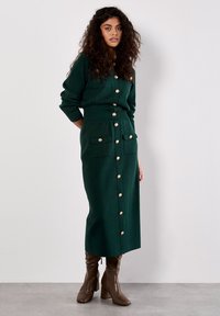 Zielona dziana sukienka z długimi rękawami, zapinana na guziki z przodu, z dwiema kieszeniami na piersi i złotymi guzikami. Noszona z brązowymi wysokimi botkami na obcasie.