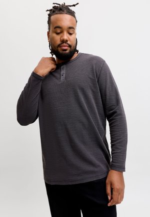 Homme aux cheveux tressés portant un t-shirt henley gris foncé à manches longues, regardant vers le bas et touchant le col avec une main.