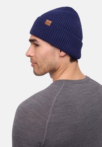Dark blue knitted beanie with a ribbed texture and a small brown leather label. Worn with a gray long-sleeve shirt.