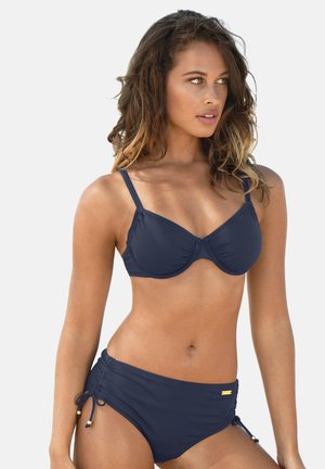 Woman posing in a navy blue two-piece swimsuit with adjustable straps and side ties against a plain light background.