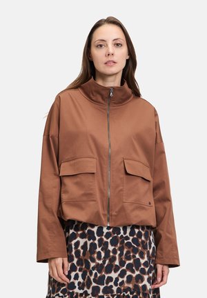 Brun, cropped jakke med høj krave, lynlås foran og to store klaplommer. Kombineret med en plettet nederdel for kontrast.